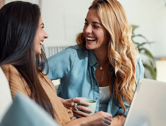 Two women are sitting on a couch, laughing together. One of them holds a mug, whilst the other has a laptop on her lap.
