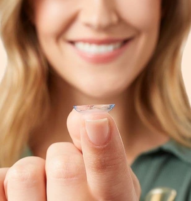 Plan rapproché sur une femme regardant une lentille posée sur son doigt avec un sourire aux lèvres. On ne voit pas ses yeux